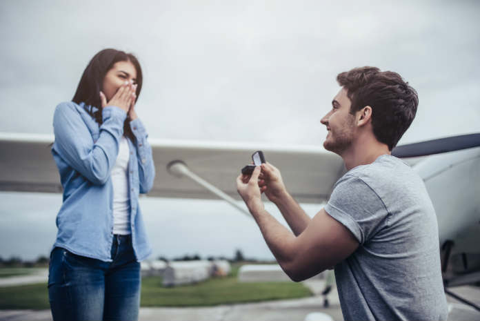 man presenting woman with a ring in front of an aeroplane
